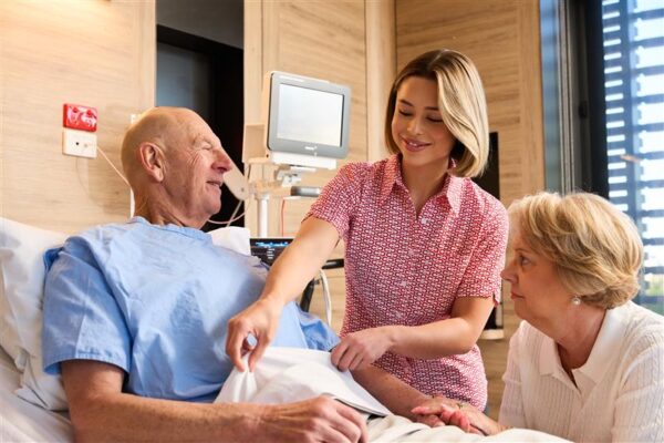 Cabrini nurse assisting a male patient in bed with their partner sitting near by